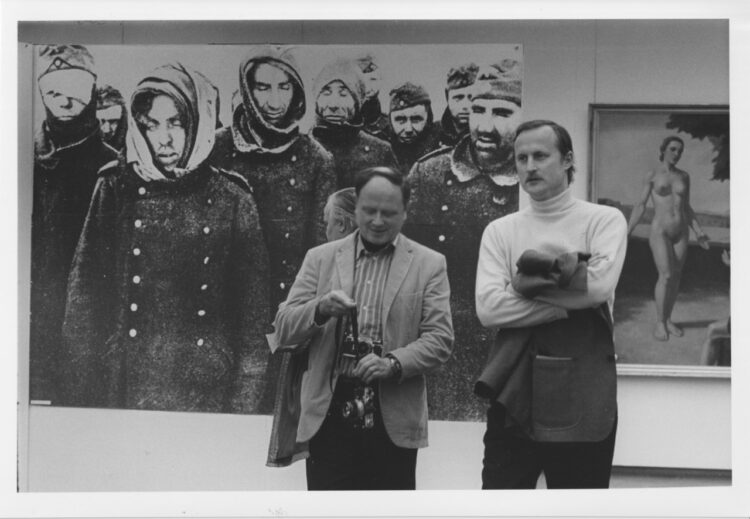 Das Foto zeigt zwei männliche Besucher der Ausstellung vor zwei in der Ausstellung zu sehende Abbildungen. Links ein Foto von Kriegsgefangenen, mit Verletzungen und darbend, reechts einen Akt im Stile der in der NS-Zeit favorisierten Malerei.