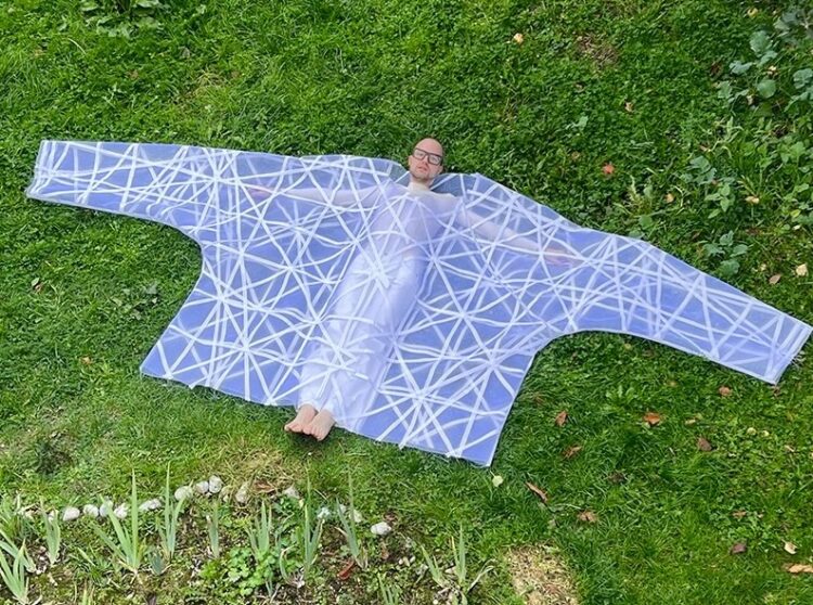 Das Bild zeigt eine Wiese. In der liegt der Künstler, eingehüllt in ein KLeidungsstück, das im sehr deutlich zu groß ist.