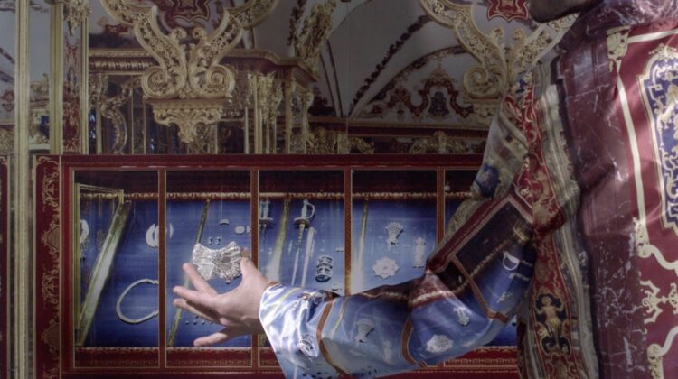 Das Foto zeigt einen Raum in püppiger, vermutlich, barocker Raumausstattung und Schmückung. Im Vordergrund ist der Arm einer nicht weiter erkennbaren Person zu sehen. Sie trägt ein Kleidungsstück, das zum Schmuck des Raumes sehr gut passt, in Blautönen, dazu weiße Manschette. In der Hand hält die Person ein silbernes Schmuckstück.